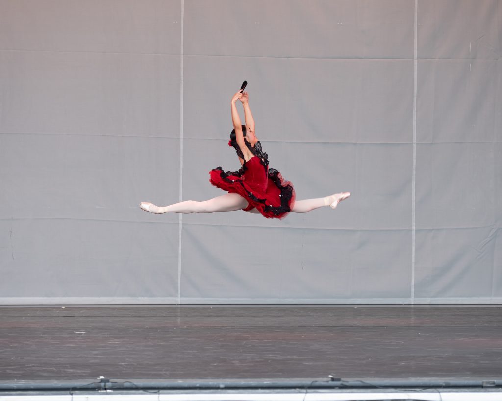 A female dancer in a red dress leaps across a stage.