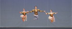 A male dancer holds hands with a female dancer on either side of him as the three leap across the stage