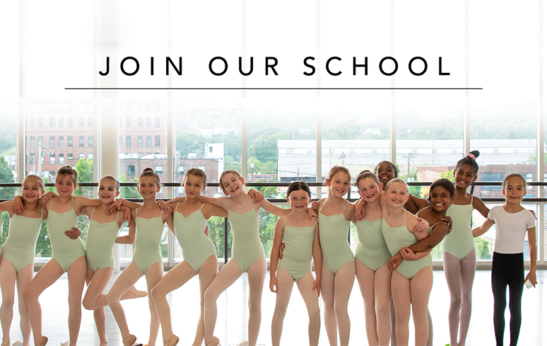 A group of students in pastel green leotards pose together with their arms over each others shoulders smiling in front of windows and ballet bars.